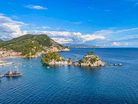 Zwei kleine Felseninseln mit grüner Vegetation in türkisfarbenem Wasser bei Parga. Ein Holzsegelboot ist sichtbar, mit Küstenstadt und Bergen im Hintergrund.