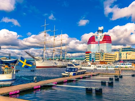 Hafen von Göteborg mit weißem Segelschiff, modernen Gebäuden mit rot-weißer Fassade, Holzstegen und schwedischer Flagge unter blauem Himmel.