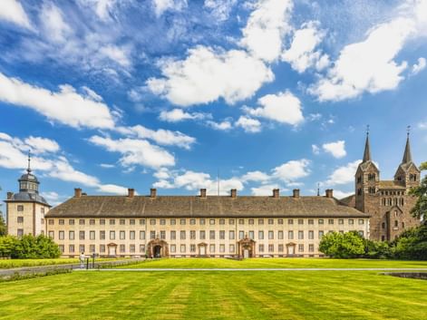 Kloster Corvey in Höxter mit langer gelber Fassade und Kirche mit zwei Türmen. Gepflegter grüner Rasen im Vordergrund unter blauem Himmel.