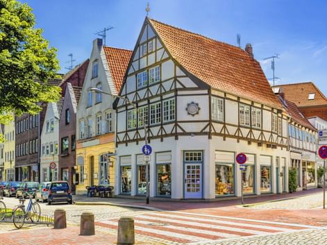 Traditionelles Fachwerkhaus an einer Ecke mit orangefarbenem Ziegeldach in Glückstadt. Die weiße Fassade zeigt dunkle Holzbalken und Geschäfte im Erdgeschoss.