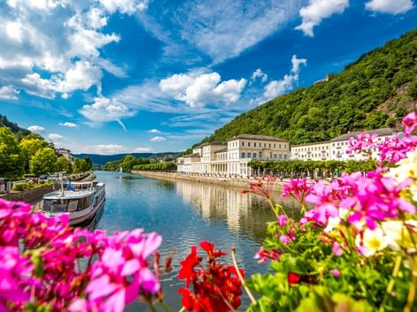 Colorful flowers frame the Lahn River in Bad Ems with historic white buildings along the waterfront, green hills, and a boat moored at the shore.