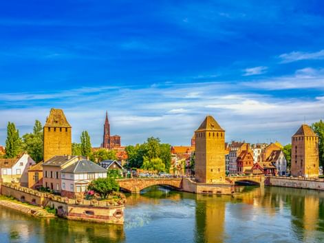 Ponts Couverts in Straßburg mit drei mittelalterlichen Türmen entlang eines Kanals, Brücke und historischen Gebäuden unter blauem Himmel.