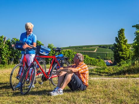 Zwei Radfahrer machen Pause im Markgräflerland. Frau steht bei Fahrrädern, Mann sitzt im Gras. Weinberge und Hügel unter blauem Himmel.