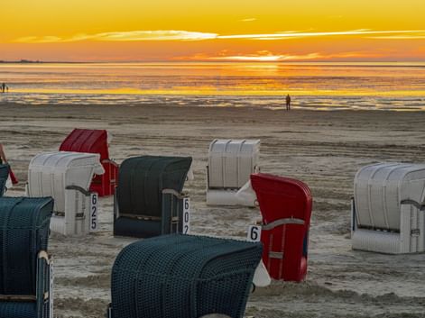 Traditionelle Strandkörbe am Strand in Bensersiel bei Sonnenuntergang. Goldener Himmel spiegelt sich im Nordseewasser mit Spaziergängern in der Ferne.