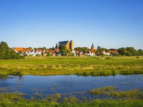 Blick auf Verden an der Aller mit historischer Kirche und Turm über weißen Häusern mit roten Dächern. Grüne Wiesen und Fluss im Vordergrund.