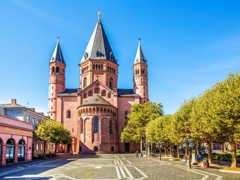 Mainzer Dom mit drei markanten Türmen und roter Sandsteinfassade auf einem sonnigen Platz mit grünen Bäumen und blauem Himmel.