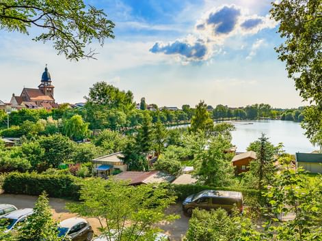 Blick durch grüne Bäume auf eine Stadt am See mit Kirchturm und historischen Gebäuden. Ein Campingplatz ist im Vordergrund sichtbar.