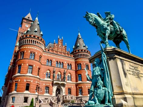 Rotes Backsteinrathaus Helsingborg mit verzierten Türmen, mit einer bronzenen Reiterstatue im Vordergrund vor klarem blauen Himmel.