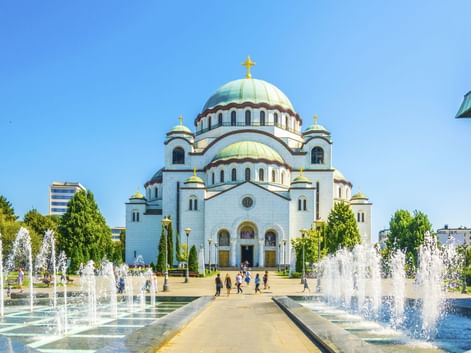Dom des Heiligen Sava in Belgrad mit weißer Fassade, grünen Kupferkuppeln und goldenen Kreuzen. Springbrunnen säumen den Weg zum Eingang.