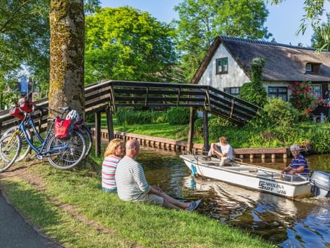 Zwei Radfahrer sitzen an einem Kanal in Giethoorn und beobachten ein Boot unter einer Holzbrücke. Fahrräder stehen in der Nähe, ein reetgedecktes Haus ist sichtbar.