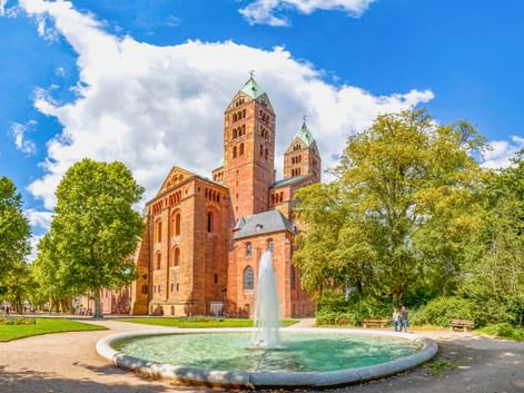Speyerer Dom mit roten Sandsteintürmen und romanischer Architektur, umgeben von grünen Bäumen. Ein runder Brunnen mit Fontäne im Vordergrund.