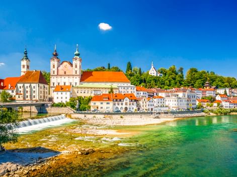 Steyrer Altstadt in Österreich mit barocken Kirchtürmen, bunten Gebäuden mit orangen Dächern entlang eines türkisfarbenen Flusses mit Wasserfall.