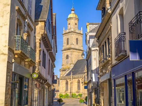 Steinerner Glockenturm der Saint-Gildas-Kirche mit goldener Kuppel durch enge Gasse in Auray. Historische Gebäude säumen beide Seiten unter blauem Himmel.