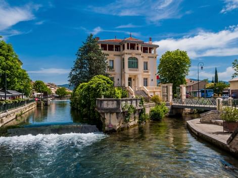 Rosa Villa mit grünen Fensterläden neben einem fließenden Kanal in L'Isle-sur-la-Sorgue, Frankreich. Ein kleiner Wasserfall und eine Brücke sind sichtbar.