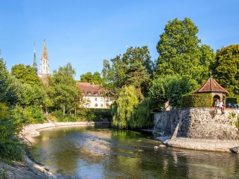 Malerischer Blick auf den Konstanzer Stadtgarten mit Fluss, Steinpavillon, üppigen Bäumen und Kirchtürmen im Hintergrund bei blauem Himmel.