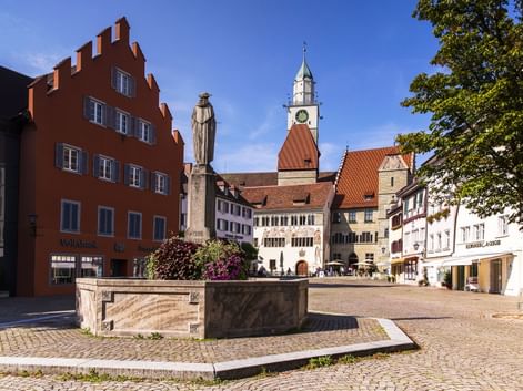 Marktplatz in Überlingen mit Steinbrunnen, rotem Giebelhaus und Kirchturm. Kopfsteinpflasterplatz mit historischen Gebäuden unter blauem Himmel.