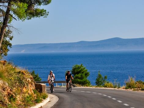 Zwei Radfahrer auf einer Küstenstraße in Dalmatien, Kroatien, mit blauer Adria und fernen Bergen unter klarem Himmel.