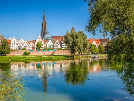 View across the Danube River to Ulm's historic old town with colorful half-timbered houses and the towering Ulm Minster spire under blue sky.