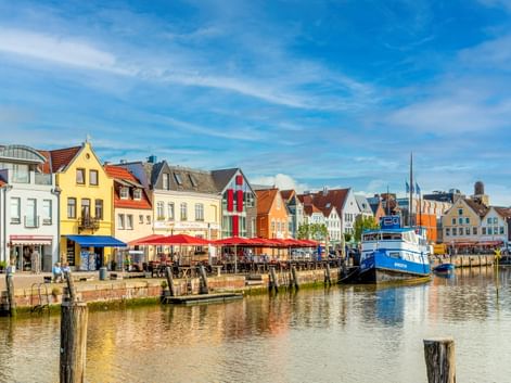 Reihe bunter historischer Häuser mit roten, gelben und weißen Fassaden entlang eines Hafenkanals. Ein blaues Boot liegt am Ufer unter blauem Himmel.