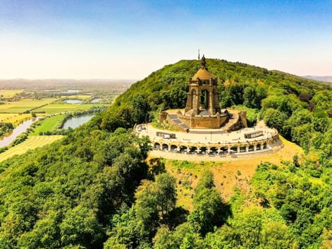 Luftaufnahme des Kaiser-Wilhelm-Denkmals auf bewaldetem Hügel in Porta Westfalica. Die Weser schlängelt sich durch grüne Felder im Tal.