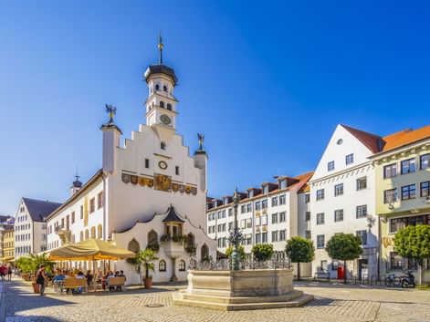Weißes Rathaus mit Glockenturm und Brunnen auf Kopfsteinpflasterplatz in Kempten. Bunte Gebäude und Straßencafés umgeben den Platz unter blauem Himmel.