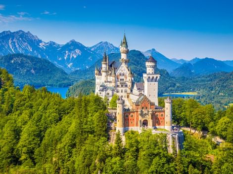 Schloss Neuschwanstein mit weißen Türmen umgeben von grünem Wald, mit Bergketten und blauen Seen im Hintergrund unter klarem Himmel.