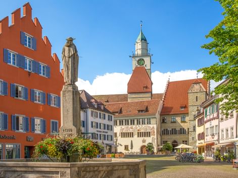Historischer Marktplatz in Überlingen mit Steinbrunnen, Statue, bunten Gebäuden und Kirchturm unter blauem Himmel.