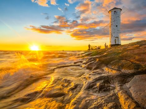 Weißer Windmühlen-Leuchtturm Mühlenbake in Swinemünde bei Sonnenuntergang. Goldene Wellen spülen über Sandstrand mit Silhouetten von Menschen.