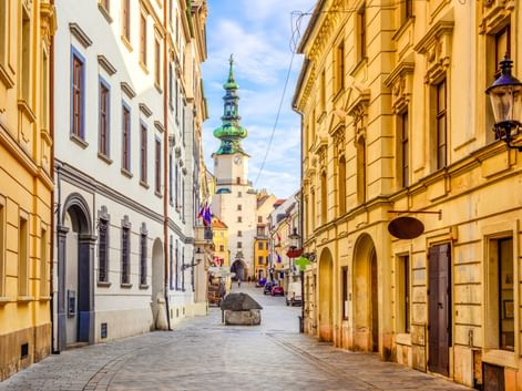 Schmale Kopfsteinpflasterstraße in Bratislavas Altstadt, gesäumt von gelben historischen Gebäuden. Ein Kirchturm mit grüner Kuppel ist im Hintergrund sichtbar.