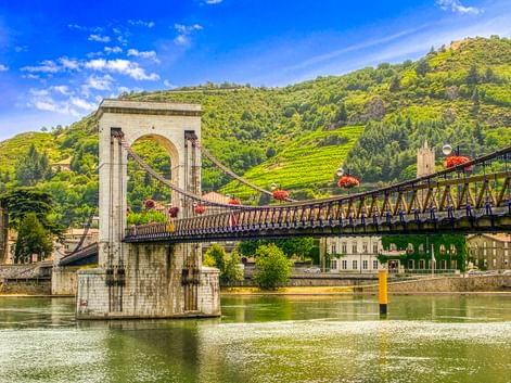 Passerelle Marc Seguin Hängebrücke über die Rhône in Tournon, mit grünen Weinbergen am Hang und historischen Gebäuden im Hintergrund.