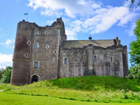 Doune Castle, eine mittelalterliche Steinfestung mit hohen Türmen und Mauern, thront auf einem grünen Hügel unter blauem Himmel mit weißen Wolken.