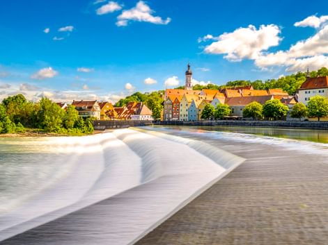 Historische Stadt Landsberg am Lech mit bunten Gebäuden und Kirchturm entlang des Lechs. Ein Wehr erzeugt fließendes Wasser im Vordergrund.