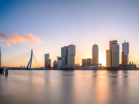 Rotterdamer Skyline bei Sonnenuntergang mit modernen Wolkenkratzern, die sich im ruhigen Wasser spiegeln. Die Erasmusbrücke links, Boote im Vordergrund.