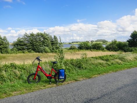 Rotes Fahrrad mit blauer Tasche auf Radweg neben Grasfeld. See und Bäume im Hintergrund unter teilweise bewölktem Himmel.
