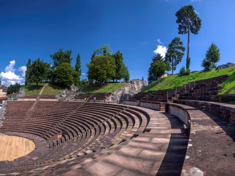 Antikes römisches Amphitheater von Augusta Raurica mit halbrunden Steinsitzreihen, grünem Hang und Bäumen unter blauem Himmel.