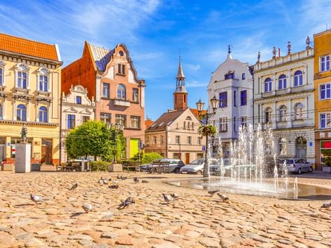 Kopfsteinpflasterplatz in der Altstadt von Tczew mit bunten historischen Gebäuden, Brunnen, Tauben und Kirchturm unter blauem Himmel.