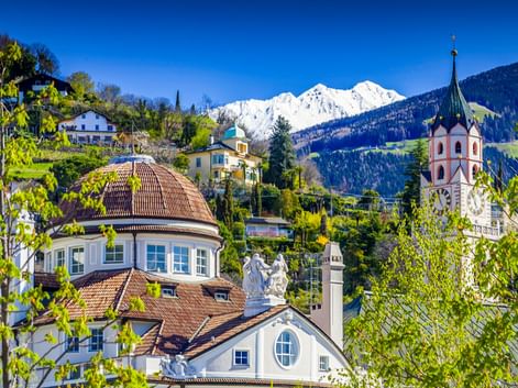 Historisches Kurhaus mit Kuppeldach und Kirchturm in Meran, umgeben von grünen Bäumen mit schneebedeckten Alpengipfeln im Hintergrund.