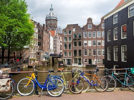 Reihe bunter Fahrräder entlang einer Amsterdamer Gracht mit traditionellen niederländischen Häusern und einem Kirchturm im Hintergrund unter bewölktem Himmel.