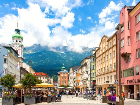 Fußgängerzone in Innsbruck mit bunten historischen Gebäuden, Straßencafés und dem Stadtturm. Alpengebirge im Hintergrund sichtbar.
