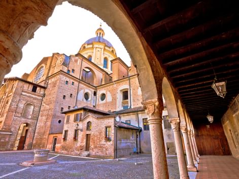 Blick durch einen Arkadenbogen auf den Dom von Mantua mit Backsteinfassade und blau gekachelter Kuppel. Das historische Gebäude wird von Steinsäulen gerahmt.