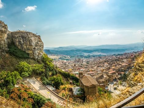 Panoramablick auf die Stadt Corleone in Sizilien mit historischen Festungsruinen auf Felsen, Terrakotta-Dächern und fernen Bergen unter blauem Himmel.