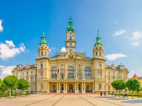 Gelbes barockes Rathaus in Győr mit drei grünen Kupfertürmen und verzierter Fassade. Menschen gehen über den Platz unter blauem Himmel.