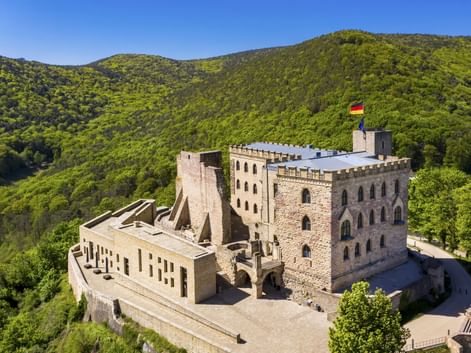 Hambacher Schloss mit deutscher Flagge auf dem Turm, umgeben von grünen bewaldeten Hügeln bei Neustadt an der Weinstraße.