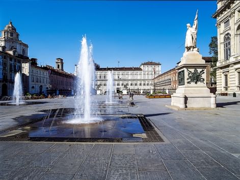 Piazza Castello in Turin mit hohen Springbrunnen im Vordergrund, weißem Denkmal rechts und historischen Palastgebäuden unter blauem Himmel.