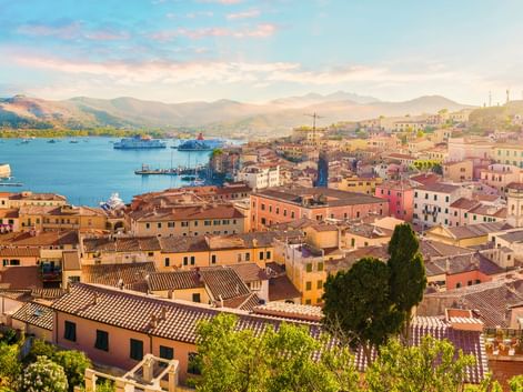 Luftaufnahme von Portoferraio mit bunten Gebäuden, Hafen mit Fähren und Bergen im Hintergrund unter weichem Abendhimmel.