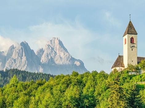 Weiße Kirche mit hohem Glockenturm in Innichen, umgeben von grünem Wald, mit dramatischen Dolomitengipfeln im Hintergrund unter blauem Himmel.