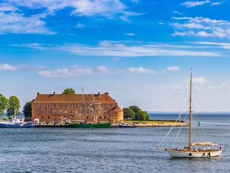 Schloss Sonderburg, ein Backsteingebäude der Renaissance, steht am Wasser mit einem Segelboot im Vordergrund und ruhigem blauen Wasser unter sonnigem Himmel.