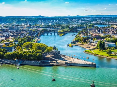 Luftaufnahme von Koblenz mit Deutsches Eck, wo zwei Flüsse zusammenfließen, Seilbahnen über dem Wasser und Festung Ehrenbreitstein im Vordergrund.