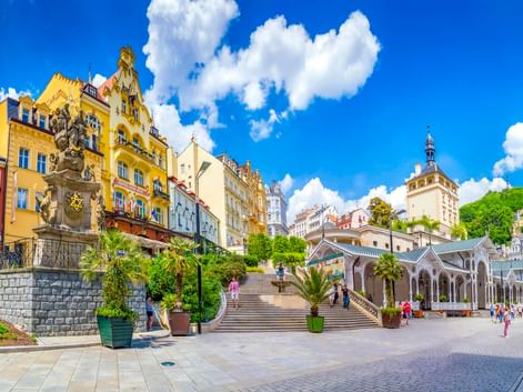 Karlsbad spa town square with colorful buildings, white colonnade, ornate fountain, and castle tower under blue sky with white clouds.