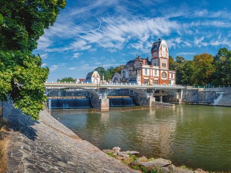 Steinbrücke über einen Fluss in Hradec Králové mit einem Uhrturm aus rotem Backstein. Grüne Bäume säumen die Flussufer unter blauem Himmel.